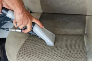 Hands using a vacuum nozzle to deeply clean a fabric car seat, removing dirt and stains.